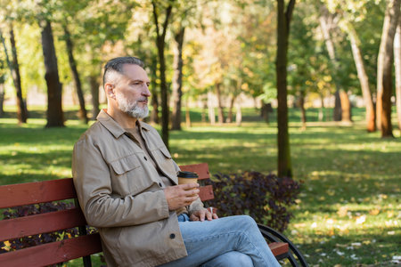 Middle aged man holding paper cup while spending time on bench in spring parkの写真素材