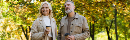 Carefree mature couple holding coffee to go while walking in park, banner
