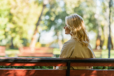 Side view of mature woman in trench coat sitting on bench in parkの写真素材