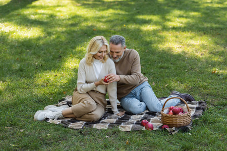 happy husband and wife holding red apple and sitting on blanket during picnic in parkの写真素材