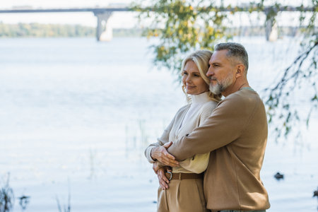 cheerful middle aged man with grey beard hugging happy blonde wife near river in parkの写真素材