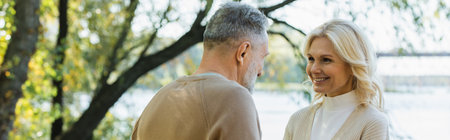 happy middle aged woman looking at bearded husband near lake in park, bannerの写真素材