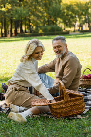 happy middle aged woman reaching food in picnic basket near cheerful husband in parkの写真素材