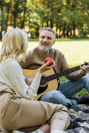joyful middle aged man with grey beard playing acoustic guitar near blonde wife with red apple during picnic in parkの写真素材