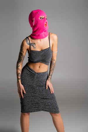 seductive tattooed woman in lurex clothes and pink balaclava looking away isolated on greyの写真素材