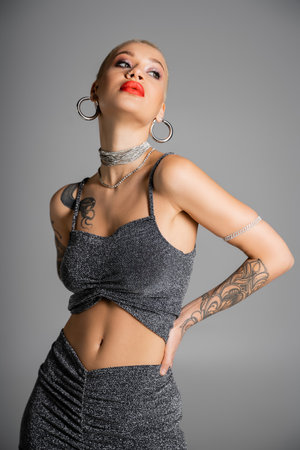 extravagant tattooed woman with hoop earrings and red lips posing with hand on waist and looking away isolated on greyの写真素材