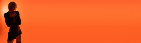 young and sexy woman in silver wig and black dress looking away on orange background with back light, bannerの写真素材