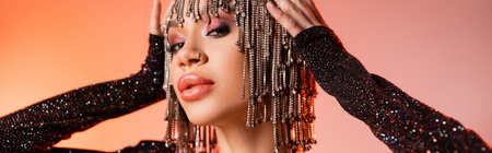 portrait of sexy young woman touching metallic wig with rhinestones and looking at camera on coral pink background, bannerの写真素材
