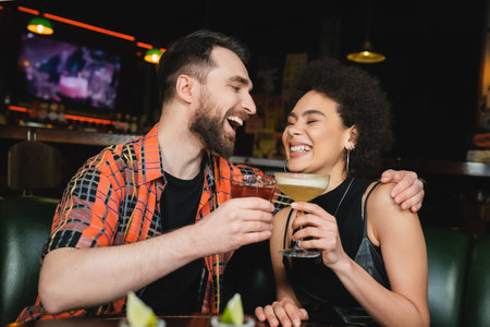 Laughing man holding negnoni cocktail and hugging african american friend near blurred tequila in barの写真素材
