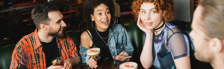 Excited interracial women holding cocktails near blurred men in bar, bannerの写真素材