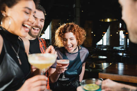 Carefree redhead woman spending time with interracial friends holding cocktails in barの写真素材