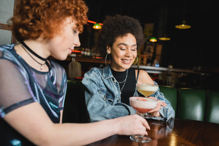 Smiling african american woman looking at pisco sour cocktail near friend in barの写真素材