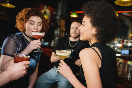 Positive curly african american woman holding pisco sour cocktail near friends in barの写真素材