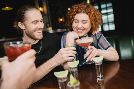 Smiling friends clinking different cocktails near tequila shots with lime in barの写真素材
