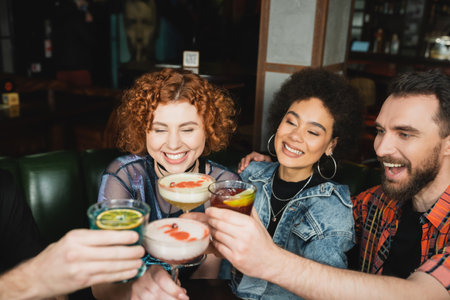 positive interracial friends smiling and toasting with different cocktails in barの写真素材