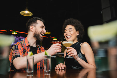 Overjoyed interracial friends holding cocktails near tequila shots on stand in barの写真素材