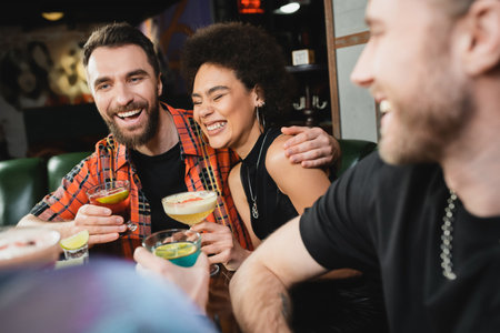 Cheerful man hugging curly african american friend with cocktail in barの写真素材