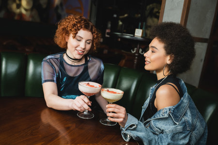 Redhead woman holding clover club cocktail and talking to african american friend in barの写真素材