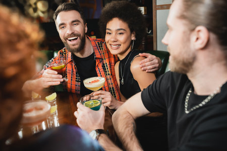 Positive multiethnic people holding different cocktails near blurred friends in barの写真素材
