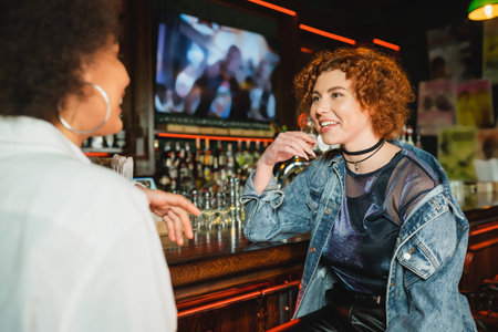 Young redhead woman talking to blurred african american friend near stand in barの写真素材