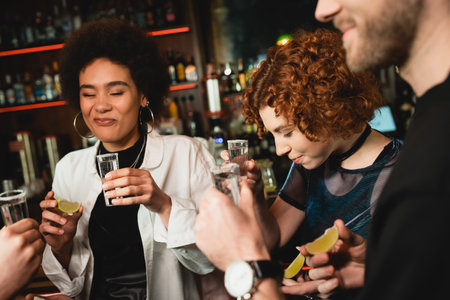 african american woman grimacing after tasting sour lime near multiethnic friends holding tequila shots in barの写真素材