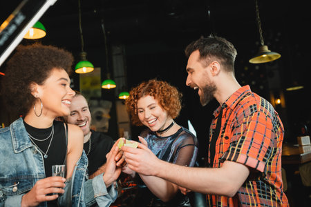 Cheerful friends holding lime and tequila near african american friend in barの写真素材