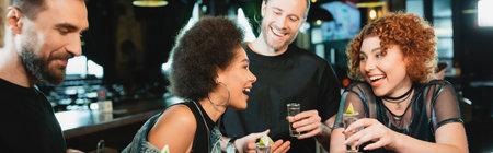 Positive interracial friends talking and holding tequila shots in bar, bannerの写真素材