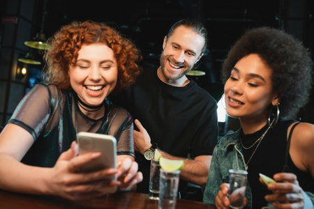 Cheerful multiethnic friends using smartphone near tequila shots in barの写真素材