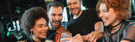 Cheerful interracial friends holding tequila shots with salt in bar, bannerの写真素材