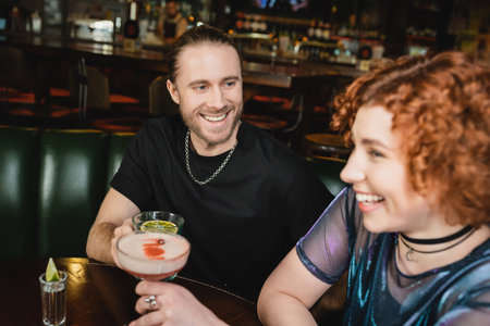 Cheerful man holding blue lagoon cocktail near blurred redhead friend in barの写真素材