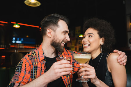 Cheerful man holding cocktail and hugging curly african american friend in barの写真素材