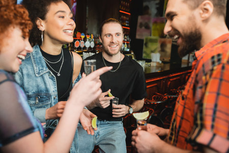 Cheerful interracial friends with tequila shots talking in bar at nightの写真素材