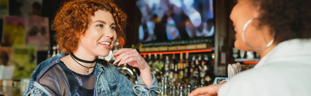 Cheerful redhead woman talking to blurred african american friend in bar, bannerの写真素材