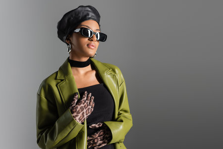 Fashionable african american woman in sunglasses and green coat standing isolated on greyの写真素材