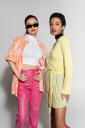 Trendy interracial women in spring outfit looking at camera on grey background
