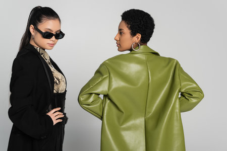 Trendy asian woman in sunglasses looking at camera near african american friend isolated on greyの写真素材