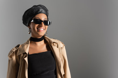 Smiling african american woman in beret and trench coat standing isolated on grey