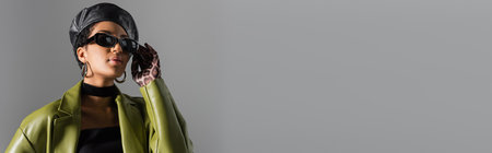 Stylish african american woman in leather beret touching sunglasses isolated on grey, bannerの写真素材