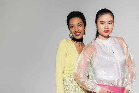 Overjoyed african american woman looking at camera near stylish asian friend isolated on greyの写真素材