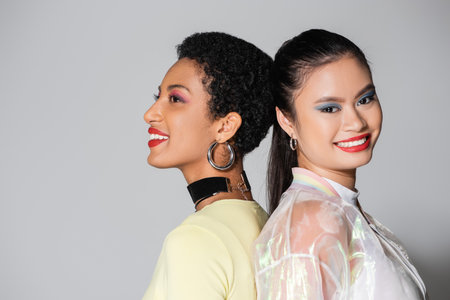 Pretty smiling interracial models with red lips standing back to back on grey backgroundの写真素材