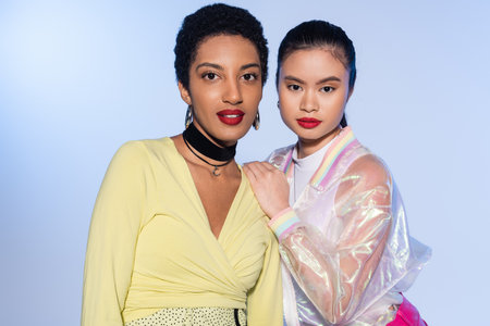 Stylish asian woman posing near african american friend isolated on blueの写真素材