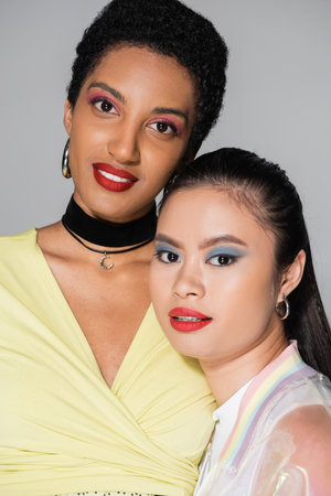 Positive multiethnic girlfriends with bright makeup looking at camera isolated on greyの写真素材