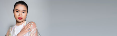 Trendy asian woman with visage looking at camera on grey background, bannerの写真素材