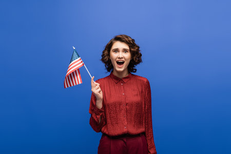 thrilled woman with open mouth holding usa flag and looking at camera isolated on blueの写真素材