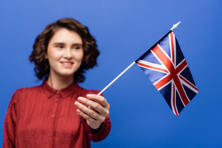 joyful student with curly hair looking at flag of United Kingdom isolated on blueの写真素材