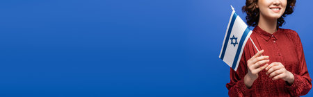 partial view of happy language teacher in red blouse holding flag of Israel isolated on blue, bannerの写真素材