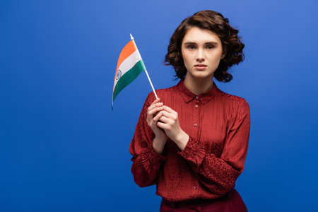 curly language teacher holding flag of India and looking at camera isolated on blueの写真素材