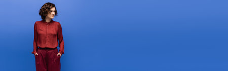 displeased language teacher in maroon color pants standing with hands in pockets isolated on blue, bannerの写真素材