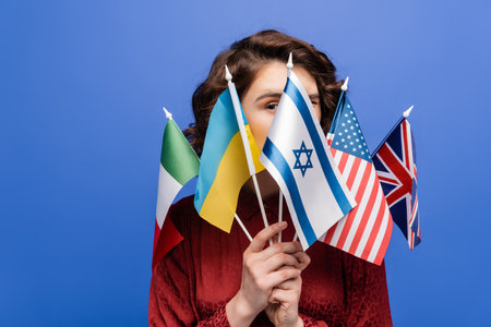 young woman looking at camera behind various international flags isolated on blueの写真素材