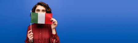 young curly woman covering face while holding flag of Italy and looking at camera isolated on blue, bannerの写真素材